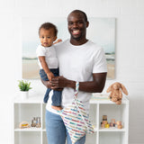 A man holding toddler and Bumkins Wet/Dry Clutch: Mickey & Friends Varsity, both in white shirts, in room with white walls and toys.