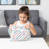 A smiling girl sits at a white table, opening a Bumkins Wet/Dry Clutch: Mickey & Friends Varsity.