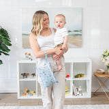 A woman holds a baby in a bright room, surrounded by toys and plants, with a Bumkins Wet/Dry Clutch: Mickey + Minnie Sunny Side Up on her wrist.