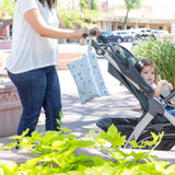 A person pushes a stroller with a child and a Bumkins Wet/Dry Clutch: Mickey + Minnie Sunny Side Up hanging from the handle.