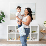 A woman cradles her baby near a toy-filled bookshelf; a Bumkins Wet/Dry Clutch: Aloha Stitch hangs from her arm.