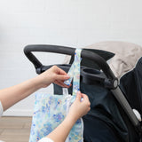 A person attaches a Bumkins Wet/Dry Clutch: Aloha Stitch to a stroller handle against a white brick wall.