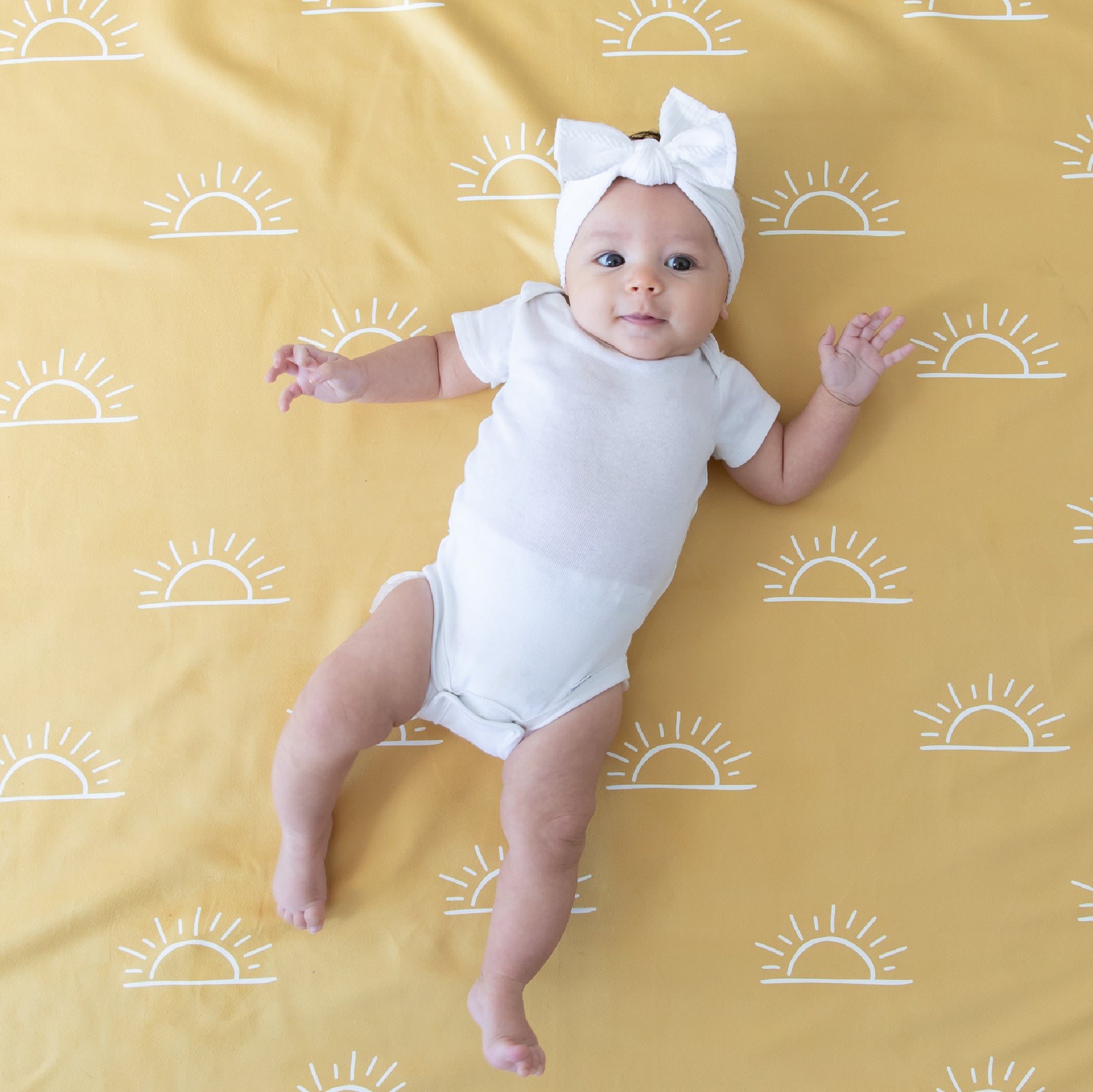 Baby in white onesie and bow headband lying on a yellow blanket with white sun patterns.