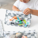 A child plays with colorful blocks in a Bumkins Clear Travel Bag 3 Pack: Charcoal Check at a white table.