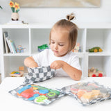 A toddler with a ponytail reaches into a Bumkins Clear Travel Bag 3 Pack: Charcoal Check at a table with colorful toys.