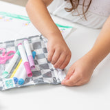 A child unzips a Bumkins Clear Travel Bag 3 Pack: Charcoal Check filled with colorful markers at a white table.