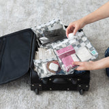 Hands placing Bumkins Clear Travel Bag 3 Pack: Charcoal Check into an open black suitcase on a carpeted floor.