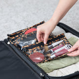 A person packs Roamio by Bumkins Clear Travel Bag 3 Pack: Leopard with makeup into an open suitcase with folded clothes on a gray carpet.