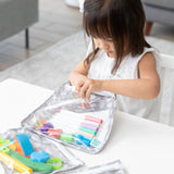 A child in a white dress opens a Bumkins Clear Travel Bag 3 Pack: Lunar Phase filled with colorful markers and scissors at a white table.
