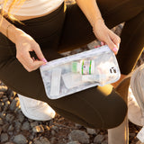A person in athletic wear kneels on rocky ground, holding a Roamio by Bumkins Clear Travel Bag 3 Pack: Disco Check with first aid supplies.