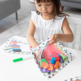 A girl in a white dress puts colorful craft supplies into the Bumkins Clear Travel Bag 3 Pack: Mickey & Friends Varsity at a table.