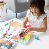 A young girl in a white dress pulls stamps out of a Bumkins Clear Travel Bag 3 Pack: Mickey & Friends Varsity on a table with art supplies.