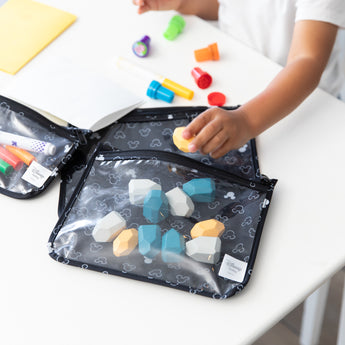 A child's hand reaches for blocks from a Bumkins Clear Travel Bag 3-Pack: Mickey Mouse Icon Black + White at a white table with art supplies.