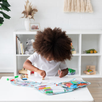 A curly-haired child draws at a white table with art supplies in Bumkins Clear Travel Bag 3 Pack: Disney Princess Ariel in a sunny room.