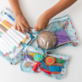 Hands opening Bumkins Clear Travel Bag 3 Pack: Disney Princess Ariel filled with markers, play dough, and toys on a white background.