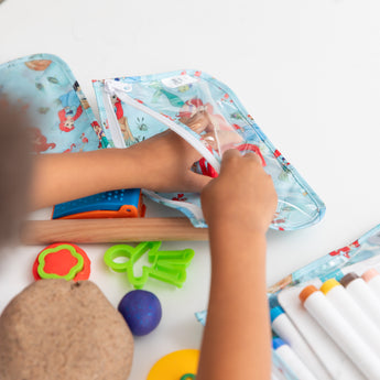 A child reaches into a Bumkins Clear Travel Bag 3 Pack: Disney Princess Ariel among art supplies scattered on a white table.