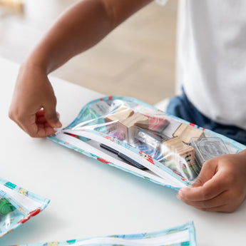 A child opens a Bumkins Clear Travel Bag 3 Pack: Disney Princess Ariel with blocks and stationery at a white table.