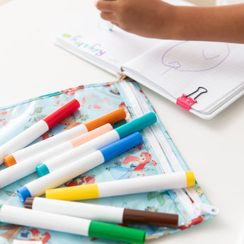 Close-up of colorful markers on top of a Bumkins Clear Travel Bag 3 Pack: Disney Princess Ariel, beside a notebook on a white table.