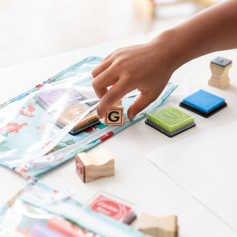 A child's hand reaches for a G block from a Bumkins Clear Travel Bag 3 Pack: Disney Princess Ariel among stamps and ink pads on a white table.