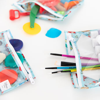 Close-up of Bumkins Clear Travel Bag 3 Pack: Disney Princess Ariel filled with art supplies on a white background.
