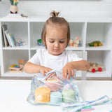 A toddler reaches into a Bumkins Clear Travel Bag 3 Pack: Aloha Stitch at a white table in front of shelves with toys and books.