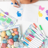 A child draws with a blue marker amid colorful markers and craft supplies in Bumkins Clear Travel Bag 3 Pack: Aloha Stitch on a white table.