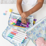 A child organizes colorful markers, glue, and pom-poms into Bumkins Clear Travel Bag 3 Pack: Aloha Stitch on a white table.