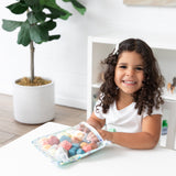 A curly-haired girl smiles at a white table, reaching into a Bumkins Clear Travel Bag 3 Pack: Aloha Stitch filled with toys.