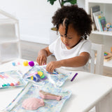 A curly-haired child at a white table reaches for art supplies stored in Bumkins Clear Travel Bag 3 Pack: Aloha Stitch.