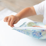 A child with a bracelet unzips a Bumkins Clear Travel Bag 3 Pack: Aloha Stitch at a white table.