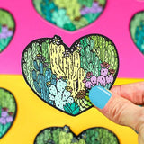 A hand holds Turtle Soups Vinyl Sticker, Cactus Love against pink and yellow surfaces adorned with similar desert-themed stickers.