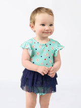 A baby smiles in a Skirt Onesie, Baseball Buddies print by Emerson and Friends, against a white background.