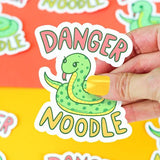 A hand holds a Turtle Soup Danger Noodle vinyl sticker of a cartoon snake against a colorful background. Scratch-resistant!.