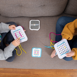 Two people sit on a gray couch, each holding a Sew Fast sewing card by Fat Brain Toy Co., a creative game challenging spatial reasoning. Colorful threads lace the cards they hold. The Sew Fast game box and another card rest between them on the wooden floor.