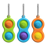 Three colorful Simpl Dimpl fidget poppers by Fat Brain Toy Co, with metal clips and tactile buttons in a pea pod shape.