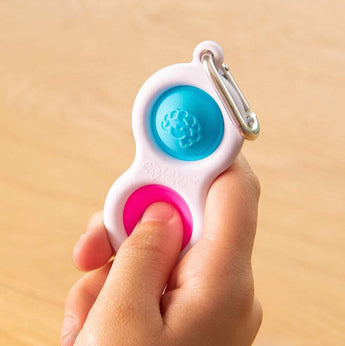 A hand holds a Simpl Dimpl by Fat Brain Toy Co, featuring silicone buttons in blue and pink on a metal clip for tactile fun.