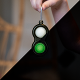 Hand holding a Fat Brain Toy Co Simpl Dimpl keychain with a glow-in-the-dark black popper featuring white and green bubbles.