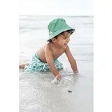 A baby in an Emerson and Friends Sea Turtles Reversible Bucket Hat plays happily in the sand by the beach.