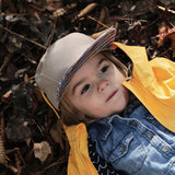 A child in a Storied Hats Lil Hat, Khaki Southwest and a yellow jacket lies on autumn leaves, gazing thoughtfully at the sky.