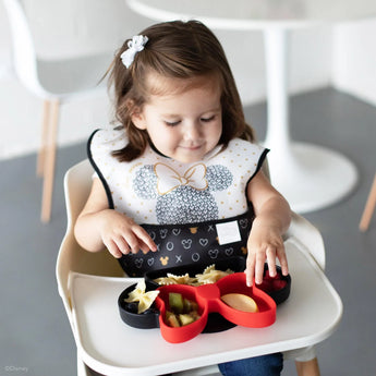 A toddler in a high chair enjoys snacks from a Minnie-shaped black & red plate, wearing a Bumkins SuperBib® 3 Pack: Love, Minnie.