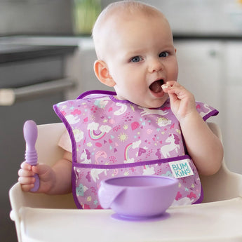 A baby in a high chair wearing a unicorn-patterned bib eats from a Bumkins Silicone First Feeding Set: Lavender.