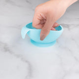 A hand holds a Bumkins Silicone First Feeding Set: Blue, demonstrating the suction cup bottom on a marble surface.
