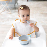 A baby with a bow and beige bib sits in a high chair, enjoying puree from a Bumkins Silicone First Feeding Set: Gray.