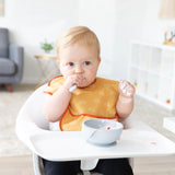A baby wearing a yellow bib in a high chair eats from a Bumkins Silicone First Feeding Set: Gray, with a chair and shelves in the background.