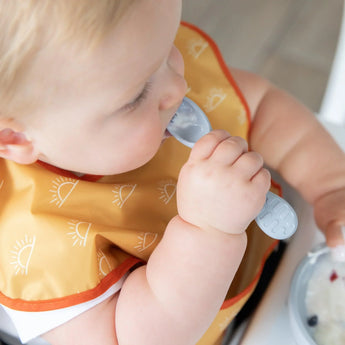Close-up of a baby wearing a yellow bib chewing the spoon of a Bumkins Silicone First Feeding Set: Gray.
