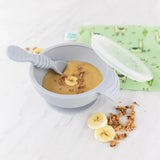 Bumkins Silicone First Feeding Set: Gray with banana puree sits among banana slices and granola and a llama print pouch on a marble surface.