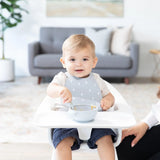A toddler in a high chair wears a gray silicone bib and eats from a Bumkins Silicone First Feeding Set: Gray with an adult nearby.