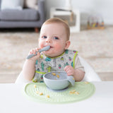 A baby wearing a cactus print bib eats from a Bumkins Silicone First Feeding Set: Gray on a green placemat in a cozy living room.