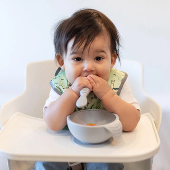A brown-haired baby sits in a high chair wearing a green bib, holding a Bumkins Silicone Dipping Spoons 3 Pack: Taffy over a gray bowl.