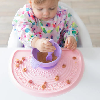 A baby in a floral bib eats from a Bumkins Silicone First Feeding Set: Lavender surrounded by cereal on a pink silicone mat.